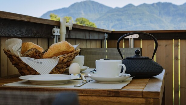 Ein Frühstück mit Brot, Eiern und Tee auf einem Holztisch, im Hintergrund ist eine Bergblick zu sehen.
