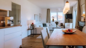 Eine moderne Küche mit Kaffeemaschine, einem Esstisch mit Obstschale, einer Couch und einem Fernseher im Wohnbereich.