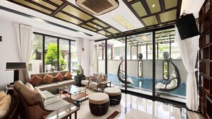 A spacious living room with a large sofa, coffee table, and ottomans. There is a large window with a view of a pool and greenery outside. - Lumiere Villa Nam Thong Phu My Hung (Ho Chi Minh City)