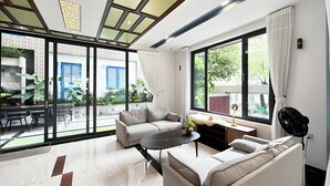 A modern living room with a sofa, a coffee table, and a fan. There is a large window with curtains, and a view of the outside. - Lumiere Villa Nam Thong Phu My Hung (Ho Chi Minh City)