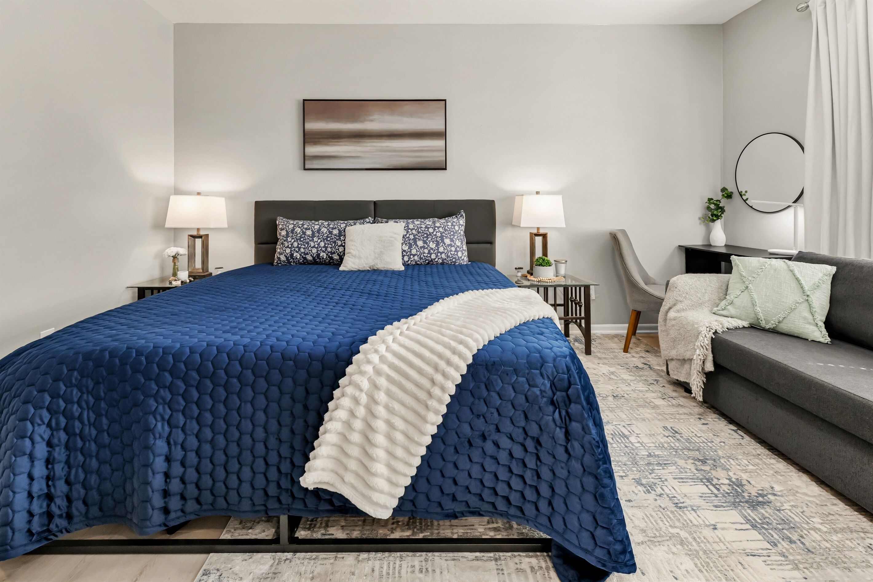 A modern bedroom with a large bed, two bedside tables with lamps, a grey sofa, and a wall-mounted mirror.