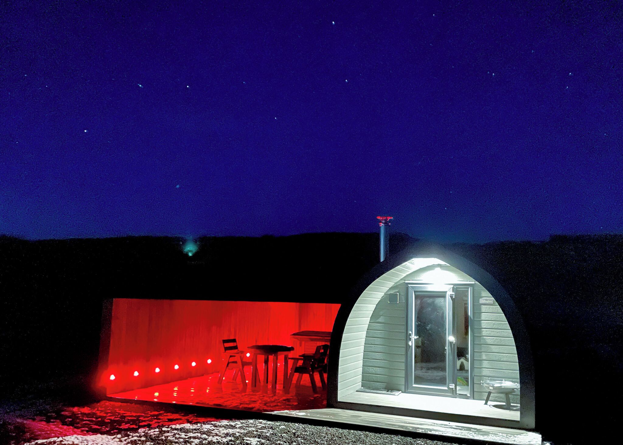 Une petite structure en forme d’arche, dotée d’une porte en verre, d’une table et de chaises à l’intérieur, se dressant sur fond de ciel nocturne.