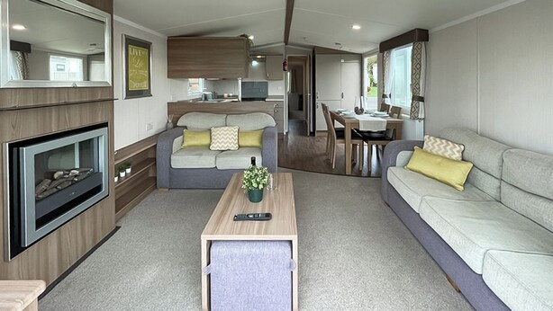 A compact living area with a sofa, a coffee table, a fireplace, and a kitchen in the background. - Birch 2 Bedroom Pet Friendly (Clynderwen)
