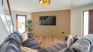 A modern living room with a large sectional sofa, a flat-screen TV mounted on a wooden panel wall, and a pendant light.