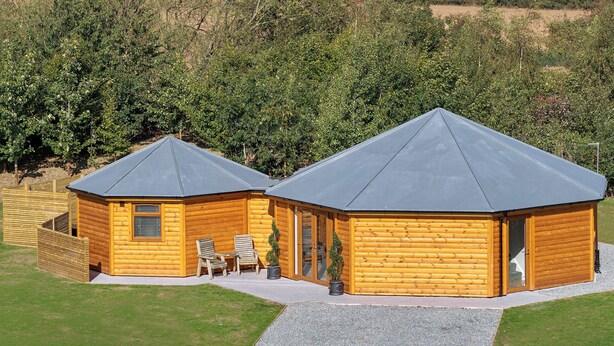 A two-story log cabin with a gravel driveway and a wooden deck with chairs.