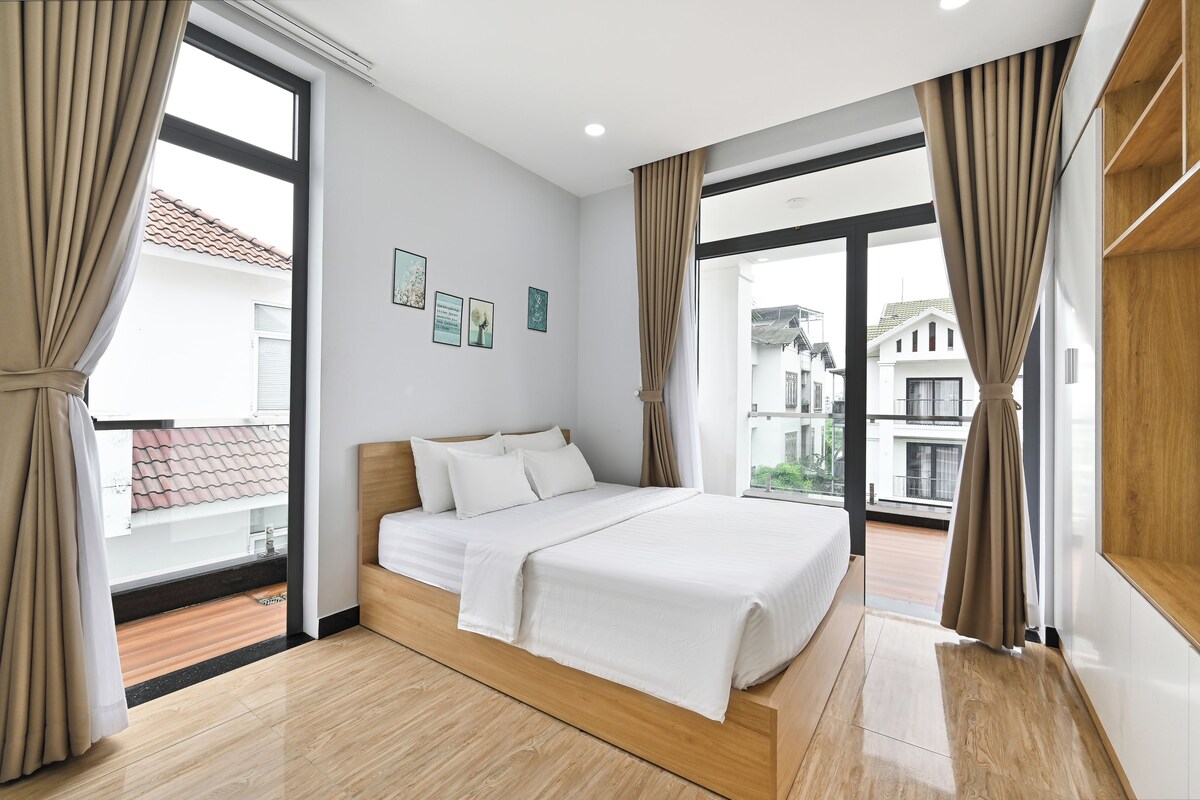 Un dormitorio con cama, ventanas amplias y vistas a edificios.