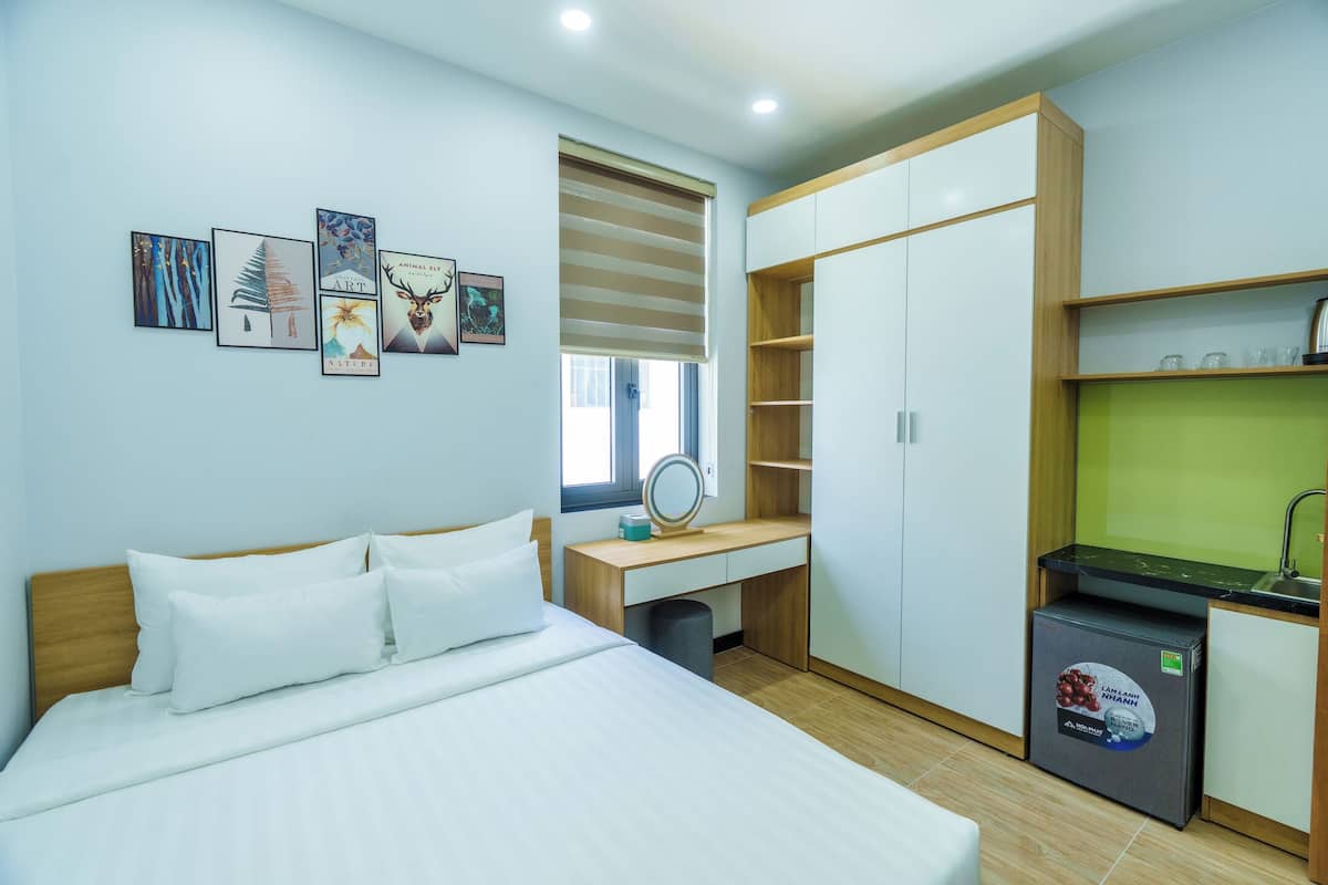 Quarto de hotel moderno com cama, escrivaninha com espelho, guarda-roupa e uma pequena cozinha equipada.