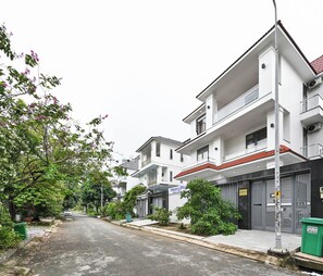 Una calle residencial con casas modernas de dos pisos, una fachada comercial visible y un contenedor verde para basura.