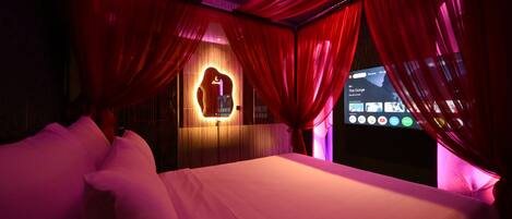 A hotel room with a bed, red curtains, and a wall-mounted TV displaying a music app.