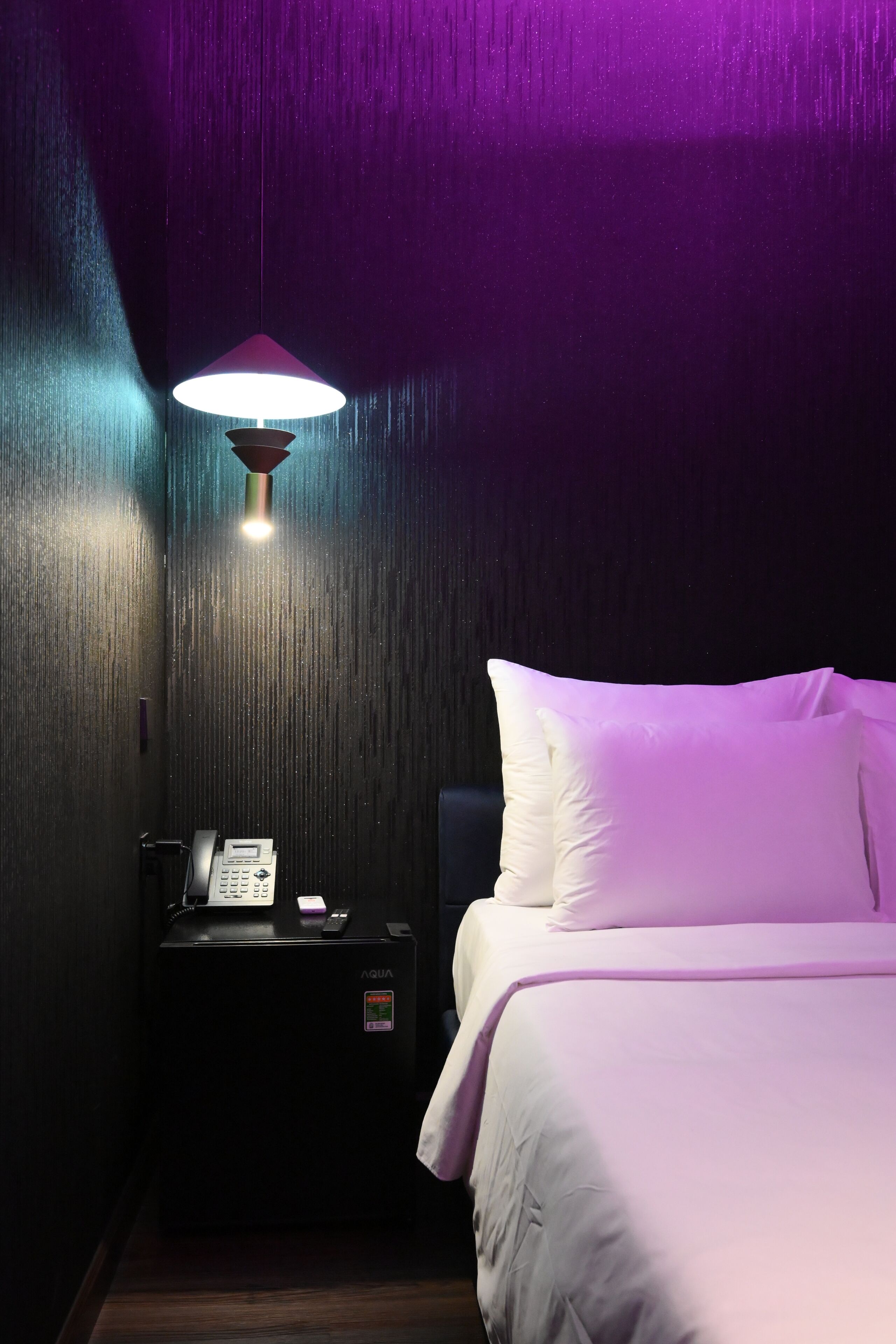 A hotel room with a bed, a nightstand with a phone, and a wall-mounted lamp.