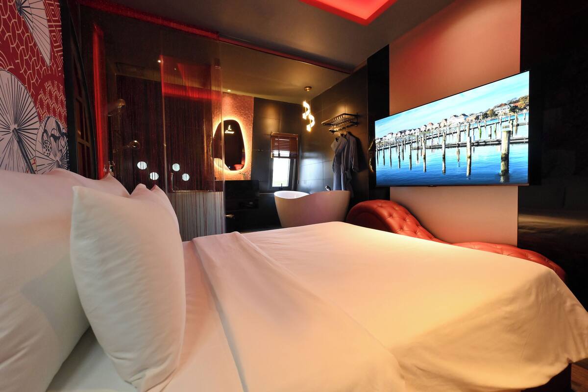 A modern hotel room with a large bed, a TV on the wall, and a view of a pier.