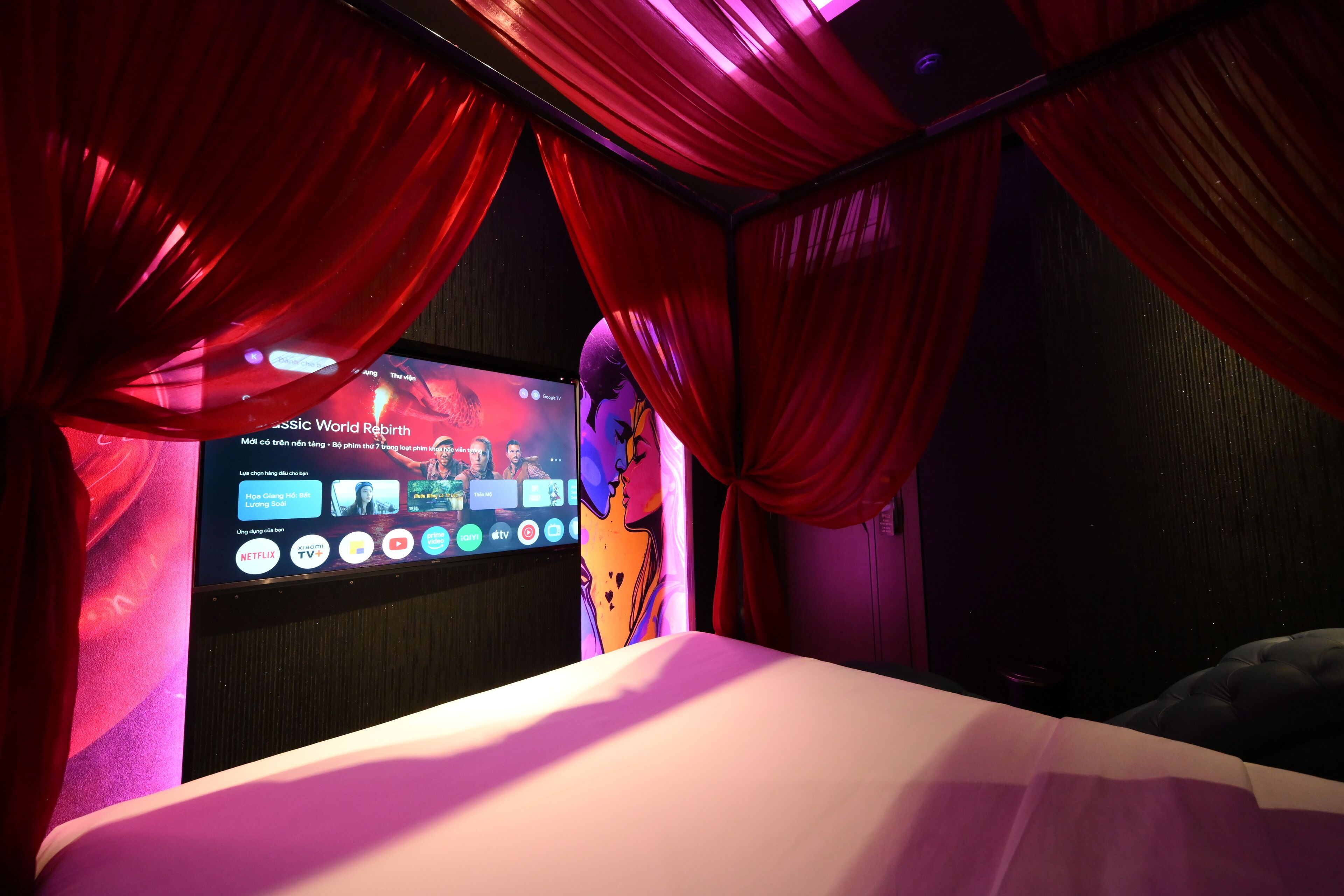 A room with a bed, red curtains, and a large screen displaying a movie.
