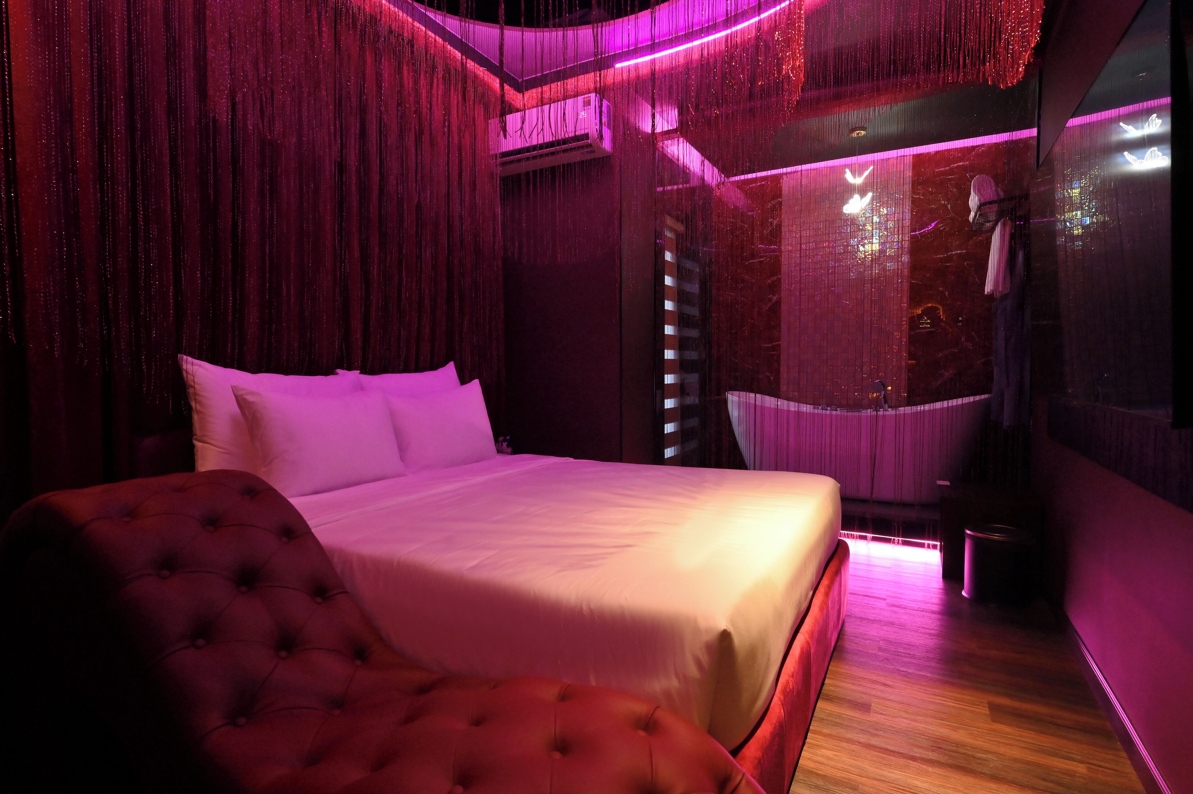 Una habitación con una cama grande, un sillón tapizado rojo y vistas de la ciudad a través de una ventana con cortinas.