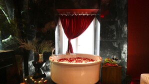 Une baignoire remplie de pétales de rose, un vase avec des fleurs séchées et un panier de fruits contenant des raisins, ainsi qu'un verre à vin posé sur une table.
