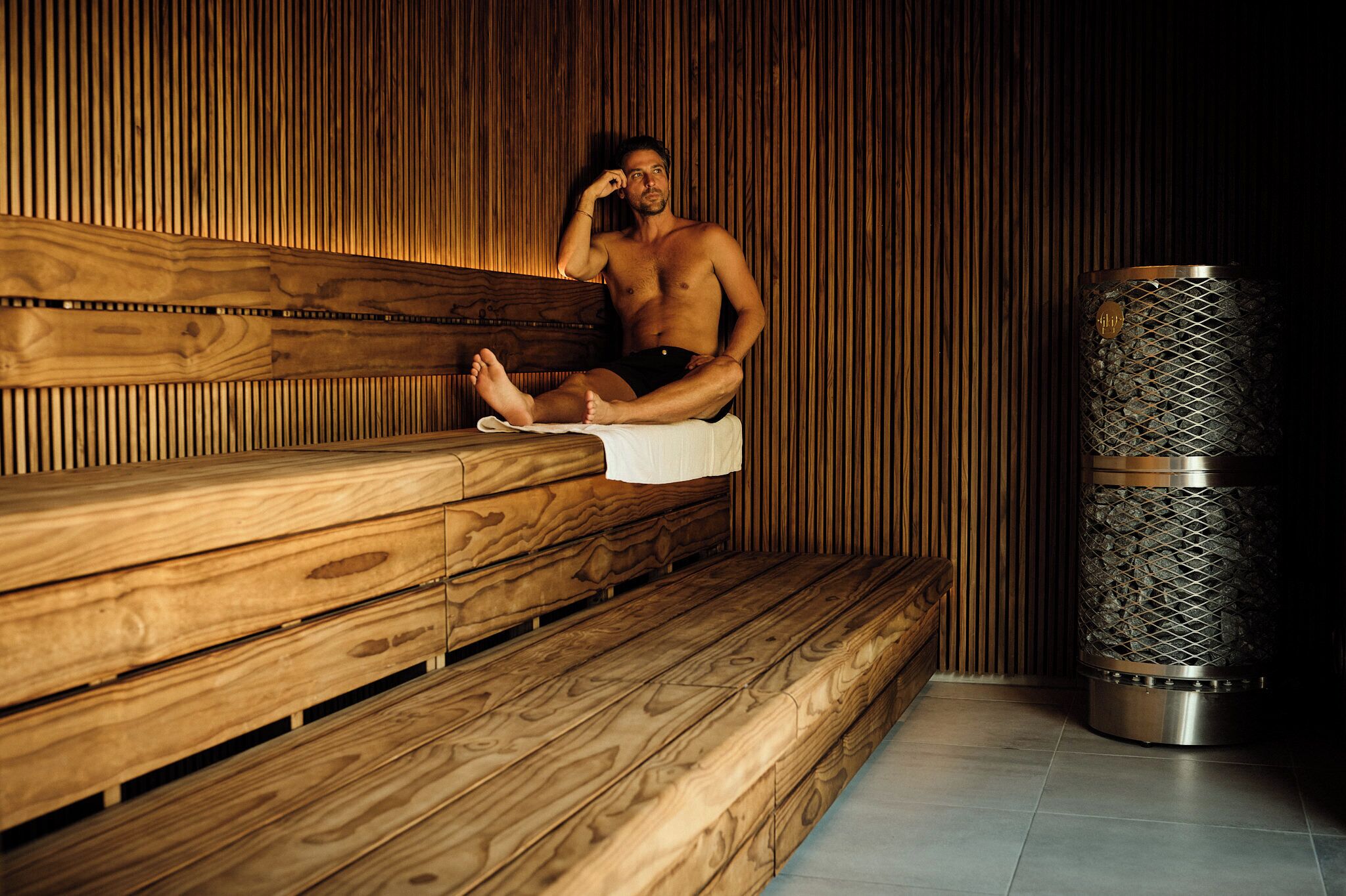 Banco de sauna de madera con una persona sentada, pared de piedra y calentador de sauna de metal.