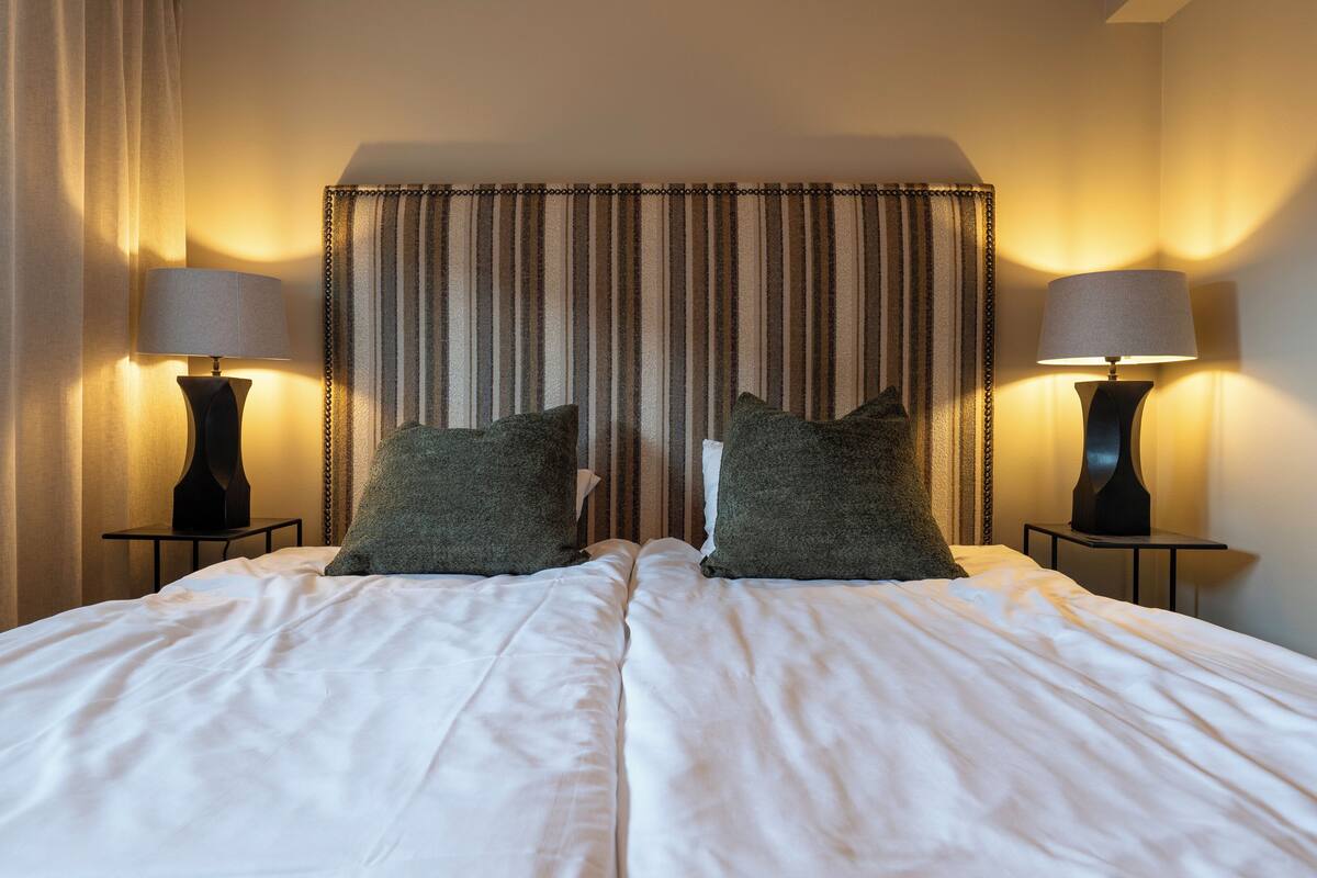 A double bed with white linens, two dark green pillows, and two bedside lamps.