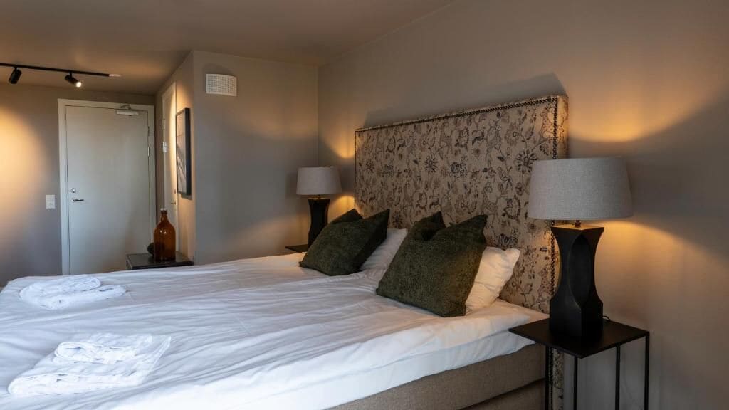 A neatly made bed with white linens and green pillows, two bedside lamps, and a floral-patterned headboard.
