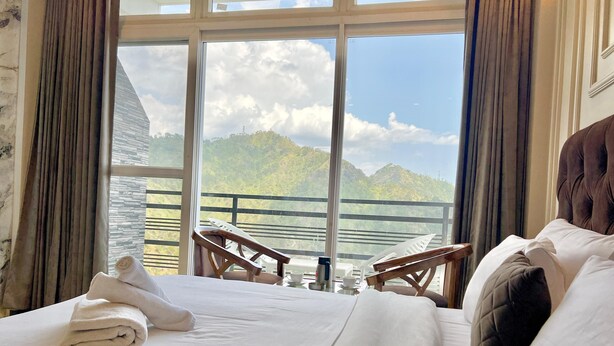 A hotel room with a bed, a view of mountains, and a balcony with chairs. - Hill Top Heaven By Elvora Hotels & Resorts (Kasauli)