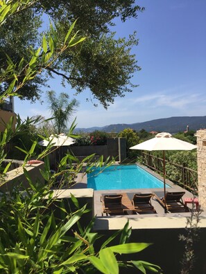 Ein Pool mit Liegestühlen und Sonnenschirmen, umgeben von Grünpflanzen und mit Blick auf eine Berglandschaft.