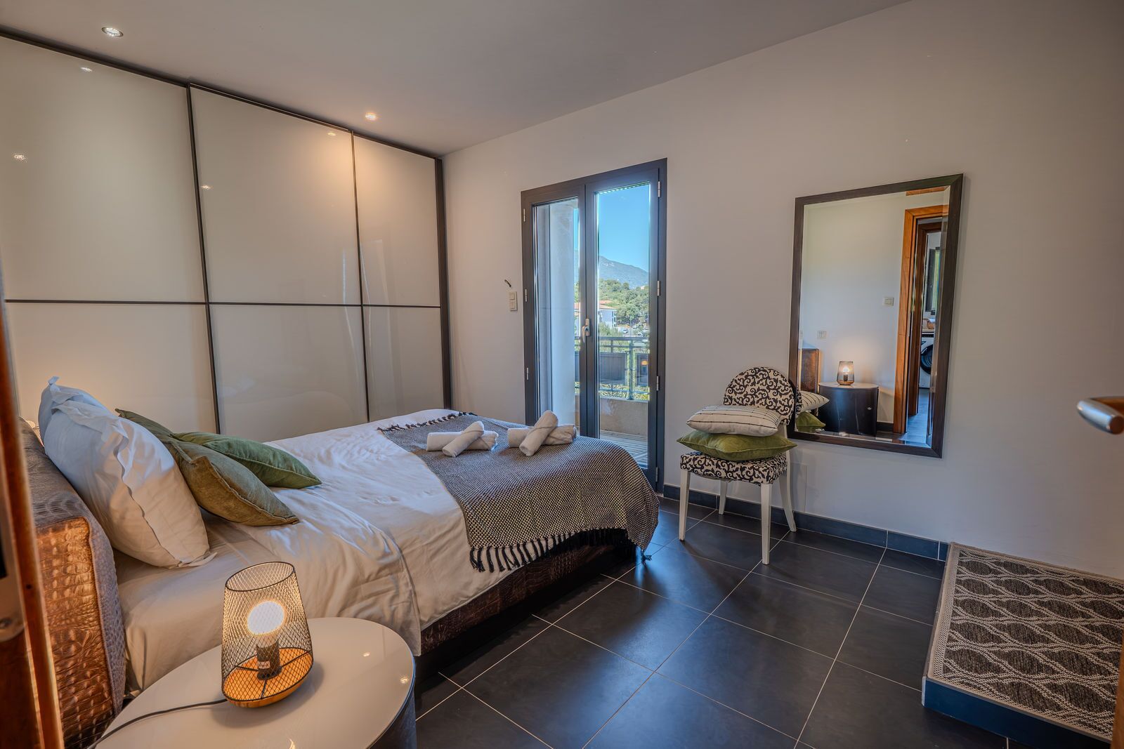 A modern bedroom with a bed, a chair, a small table, a mirror, and a view of the outdoors.