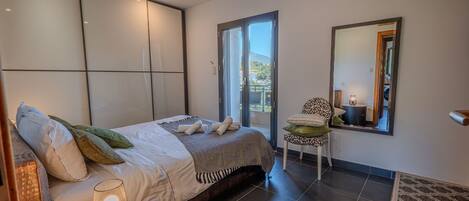 A modern bedroom with a bed, a chair, a small table, a mirror, and a view of the outdoors.