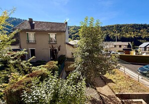 A residential area with a house featuring a sloped roof, a small garden, and a view of a forested hill in the background.
