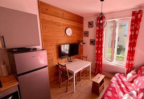 A compact room with a wooden wall, a small desk with a TV, a bed with red patterned bedding, and a window with red curtains.