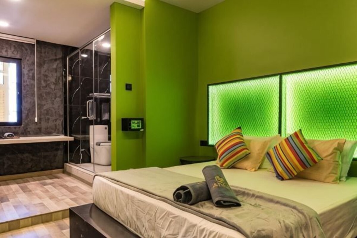 A modern bedroom with a green wall, a bed with striped pillows, a shower area, and a visible window.