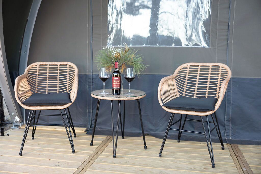 Two wicker chairs with black cushions and metal legs, a round table with a bottle of wine and two wine glasses, and a large window with a view of trees.