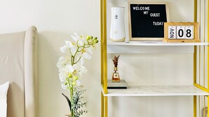 Ein Hotelzimmer mit weißem Sofa, einem kleinen runden Tisch mit Blumenvase, einem Regal mit einer Willkommensanzeige und einem Kalender.