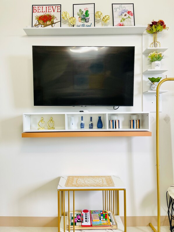 Una sala de estar con televisor montado, estanterías con objetos decorativos, una mesita con juguetes y una silla dorada.