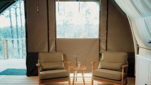 Um quarto com tenda, duas cadeiras e uma mesa pequena, com vista para árvores pela janela, e um deck de madeira lá fora.