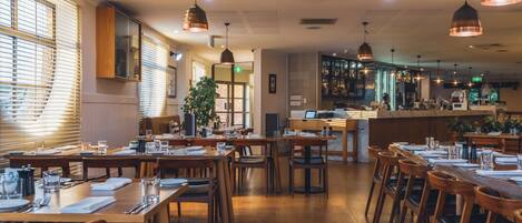 Un restaurante con mesas y sillas de madera, una zona de bar y grandes ventanales con persianas.