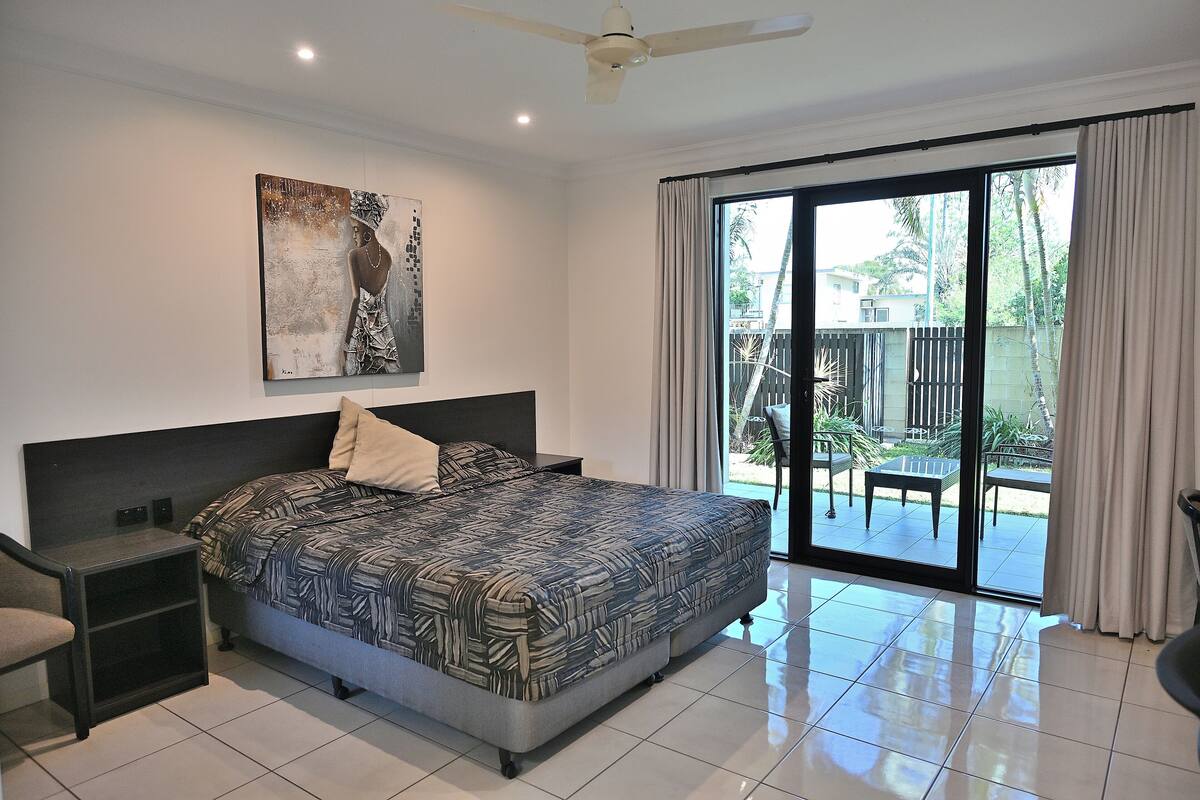 A bedroom with a large bed, a nightstand, a chair, a ceiling fan, and a sliding glass door leading to an outdoor area.