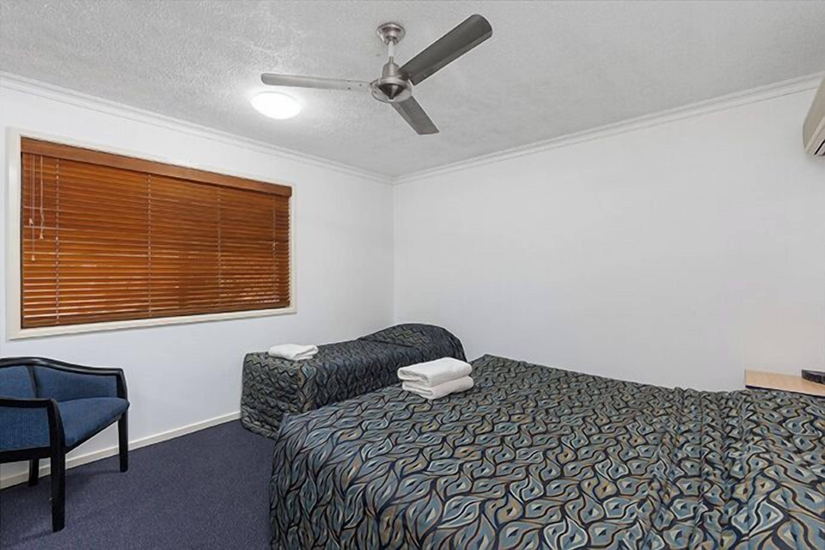 Un dormitorio con cama, una silla, ventilador de techo y ventana con persianas.