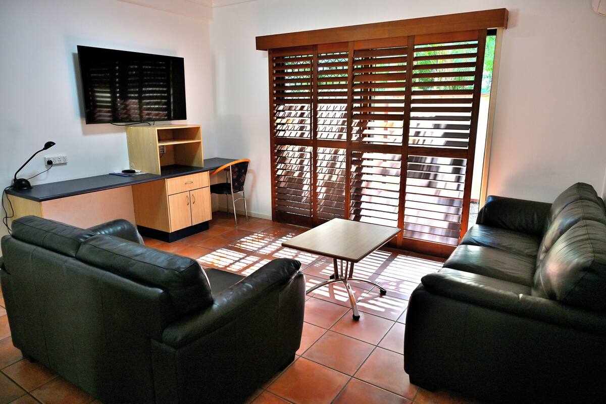A living room with a sofa, a coffee table, a TV mounted on the wall, and a window with blinds.