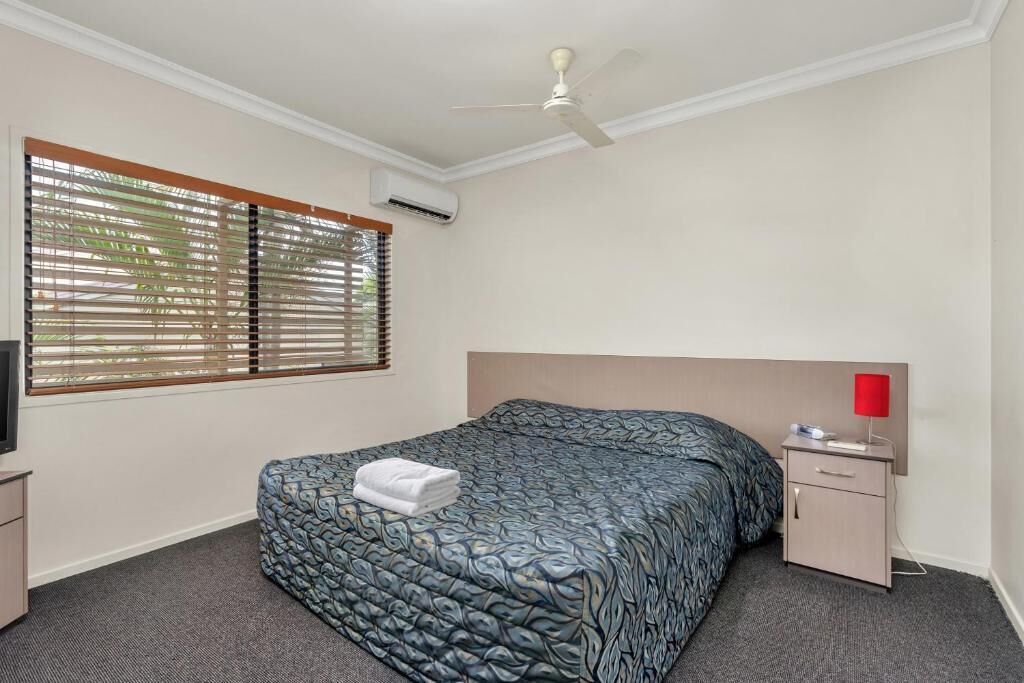 A hotel room with a bed, a nightstand with a lamp, a ceiling fan, and a window with blinds.