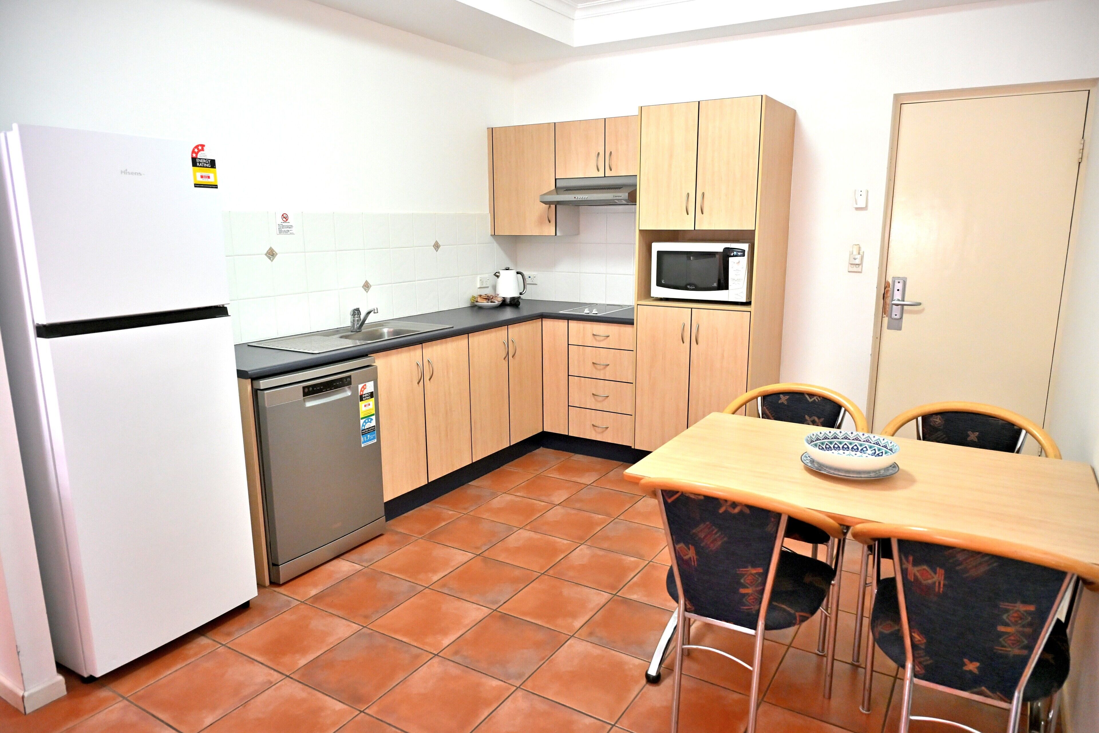 Una cocina compacta con refrigerador, microondas, lavamanos y armarios de madera. Hay una mesa de comedor con sillas y un pequeño cuenco decorativo encima.