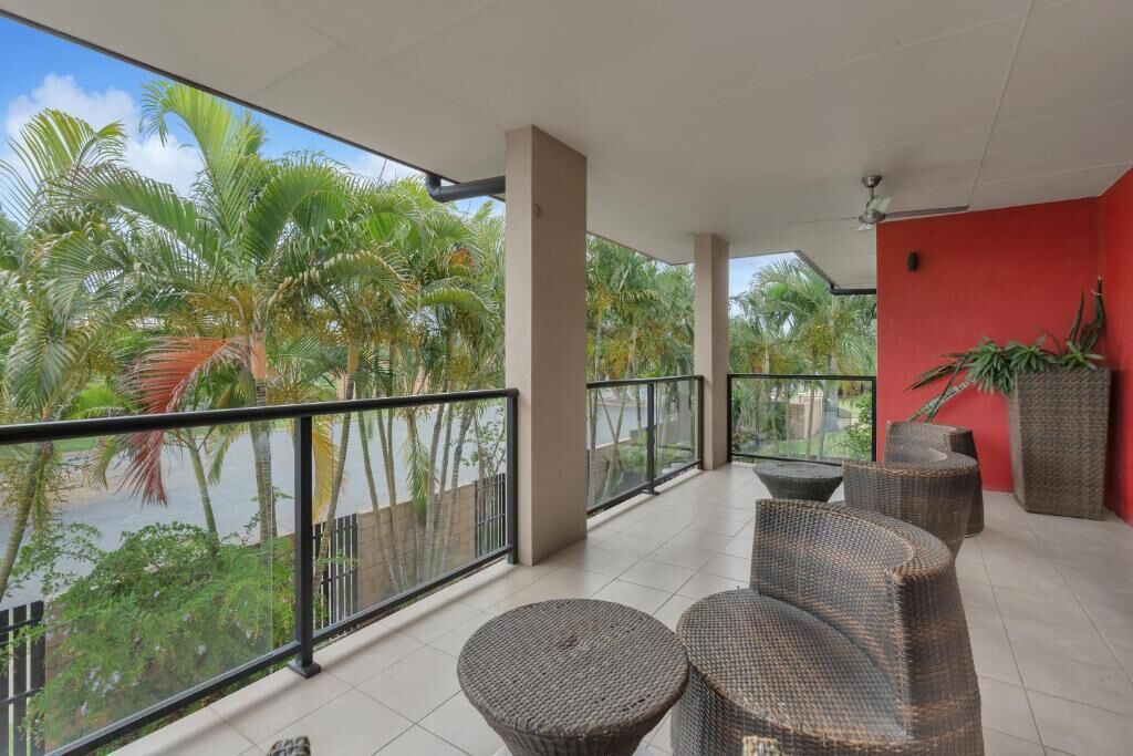 Een balkon met rieten meubilair, een rode muur en uitzicht op palmbomen en water.