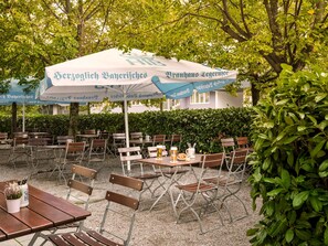 Een terras met tafels en stoelen, parasols en een haag op de achtergrond.