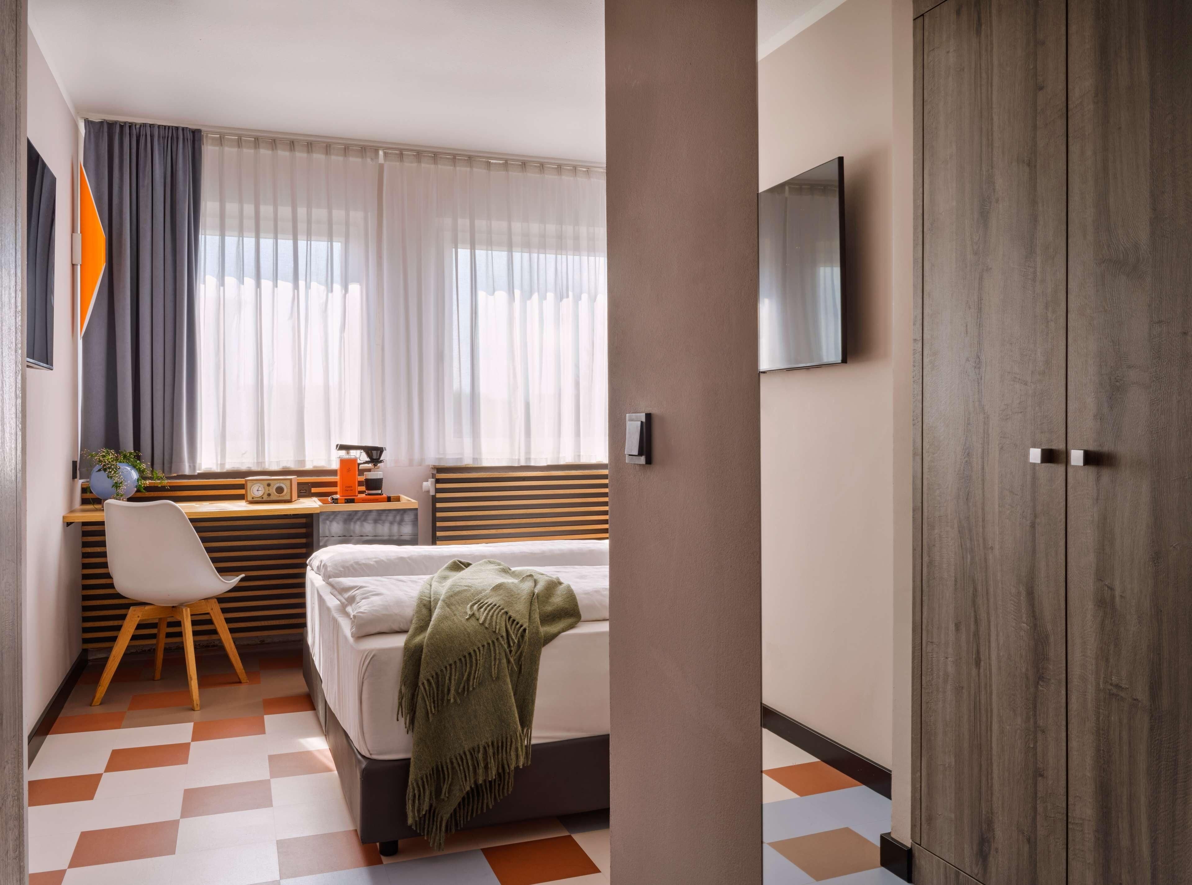 A hotel room with a bed, a desk with a chair, a television mounted on the wall, and a large wardrobe.