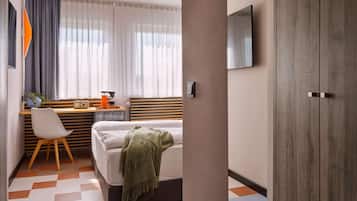 A hotel room with a bed, a desk with a chair, a television mounted on the wall, and a large wardrobe.