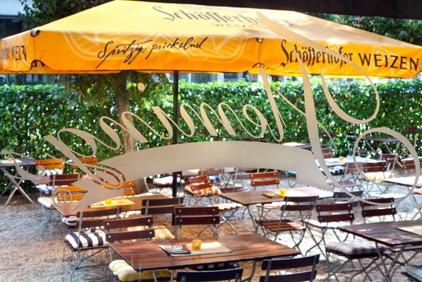 Outdoor seating with tables and chairs, an umbrella, and greenery in the background. - Flemings Hotel München-schwabing (Munich)