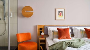 A modern hotel room with a shower, a round wooden accent wall, a bed with multiple pillows, and a small bedside table with a lamp. - Flemings Hotel München-schwabing (Munich)