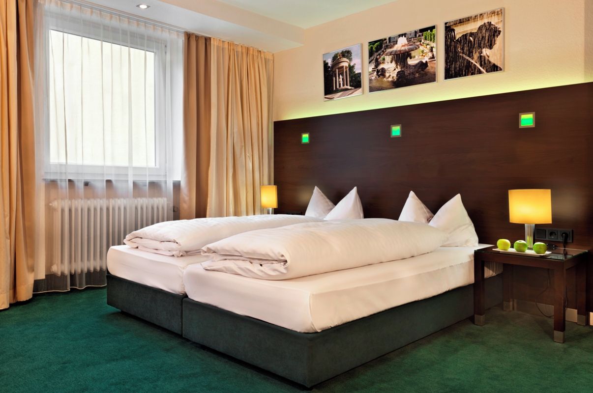 A hotel room with a large bed, two bedside lamps, a radiator, and a window with curtains.