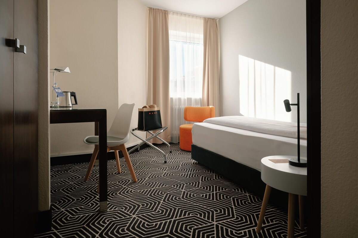 A hotel room with a bed, a desk with a chair, a small table with a lamp, a window with curtains, and a patterned floor.