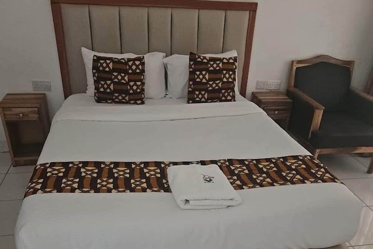 A neatly made bed with patterned bedspreads and pillows, a wooden nightstand, a chair, and a pair of slippers on a rug.