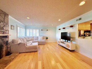 Sala de estar con sofá modular, televisor de pantalla plana, piso de madera, chimenea de piedra y una cocina visible a través de un panel de vidrio.