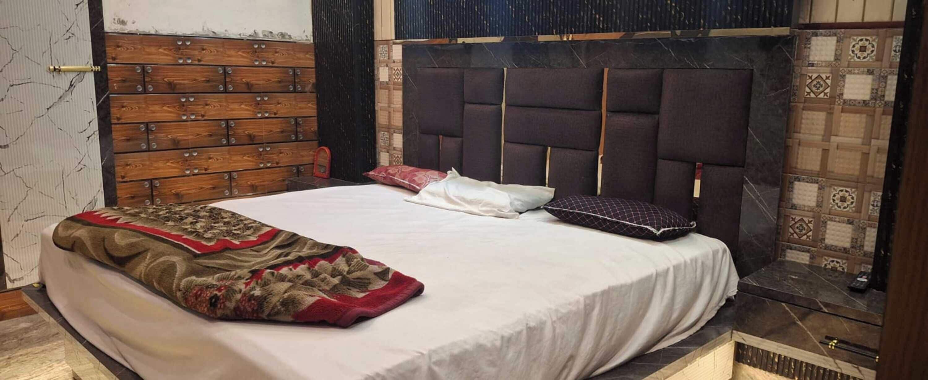 Ein Bett mit weißer Bettdecke, einem gemusterten Überwurf und zwei Kissen. Das Kopfteil ist dunkel und mit einer gepolsterten Struktur versehen.