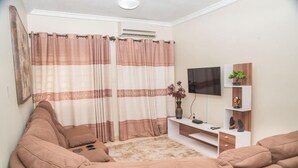 A living room with a beige sofa, a TV on a white stand, and a decorative plant. - Regimanuel Estates  (Kwabenya)